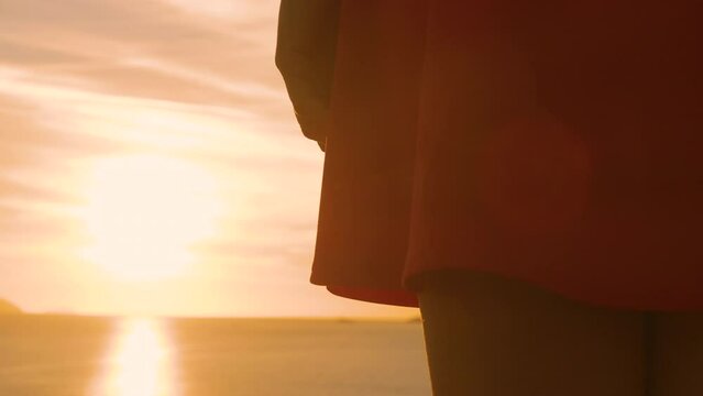 LENS FLARE, CLOSE UP: Rays of setting sun peek over hem of her red summer dress. Young lady is standing at the seashore and watching beautiful golden sunset. Calm evening moments by the Adriatic Sea.