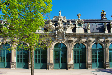 Facade of ancient building with beige walls, statues, decorative elements, windows with arches and green frames. Baroque style. Green tree and blue sky. Dresden, Art Gallery, Germany, May 2023 