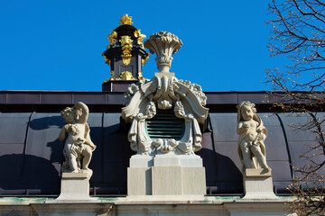 Sculptures of small antique boys on pedestal and coat of arms with floral decoration on the roof of old building with brown roof. Baroque style. Blue sky. Dresden, Art Gallery, Germany, May 2023
