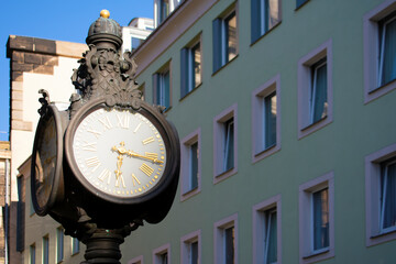 Large antique street clock with four sides in metal frame with decorative elements and golden arrows. Facade of the building with beige walls and windows in the background. Dresden, Germany, May 2023 - Powered by Adobe