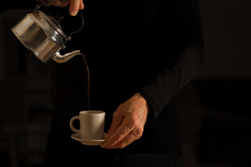the hands of an unrecognizable man in the shadows pouring a cup of coffee from a metal teapot