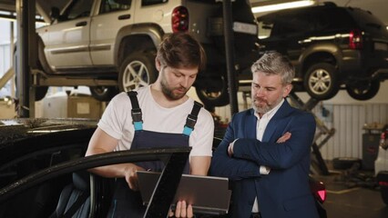 Auto mechanic showing data on the computer for mature haired male customer. Gray-haired male client communicates with an electrician in a car service. Car service, repair, maintenance clients