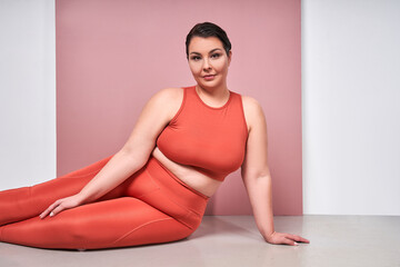 Fototapeta na wymiar Smiling plus size woman in fitness wear posing at the studio with pink background