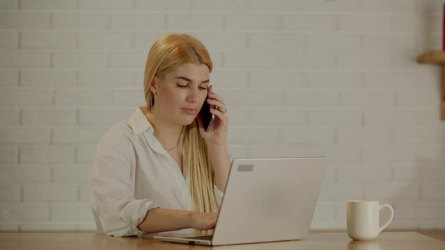 Young businesswoman working at home, sitting by workdesk with laptop, having phone conversation. Successful freelancer