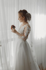 Preparation for the wedding. Beautiful young bride in white wedding dress indoors. Luxury model looks at wedding shoes, at home in studio room with big window. The girl shows
