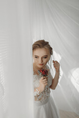 Preparation for the wedding. Beautiful young bride in white wedding dress indoors. Luxury model looks at wedding shoes, at home in studio room with big window. The girl shows
