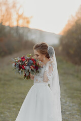 Wedding photo in nature. The bride is standing in the forest. The bride in a beautiful dress with a long train, holding her bouquet of different roses, smiling sincerely at the camera. Portrait