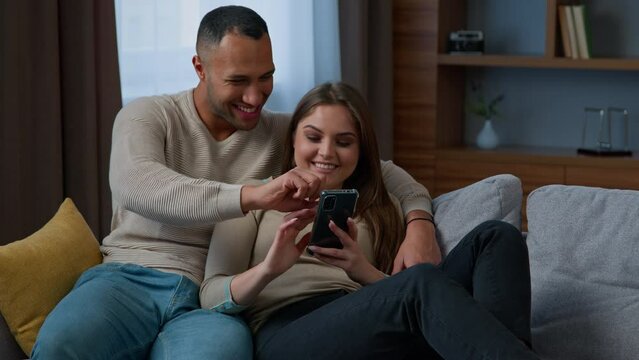 Multiracial couple African American man husband Caucasian woman wife rest on couch together multiethnic happy homeowners use mobile phone shopping online make internet order buy home goods laughing