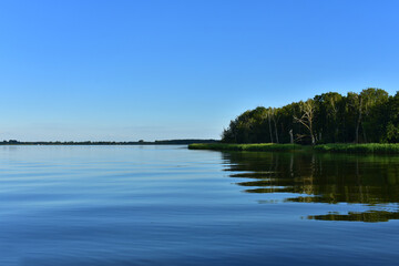 Beautiful landscape with blue lake, an island with dark green trees and green grass, and horizon with a forest. Quiet calm water. Sunny summer day with a blue sky. Dziwnów, Poland, Baltic, June 2022 