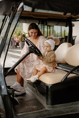 Adorable child in a funny white cap dress and sweater driving a golf cart. Mom is touched by the child sitting next to him in the car. Happy weekend