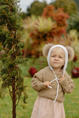 Adorable child, dressed in a funny white hat, sweater, and dress, explores the park with playful enthusiasm, while attempting to remove the hat.