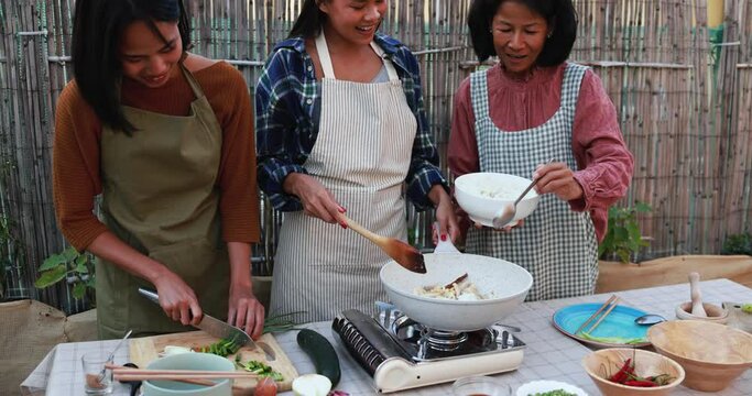 Happy asian family preparing together traditional thai rice food at house terrace - Mother and adult daughters cutting and adding fresh ingredients to the cooking pan