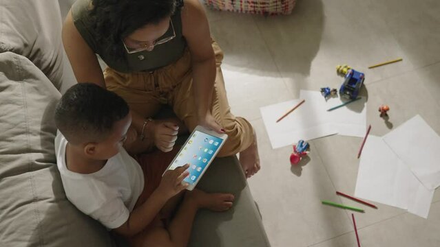 Latina mother cute little preschool son at home play on wireless tablet device. Smiling mom small kid boy look at pad screen have fun together use modern electronic tech concept
