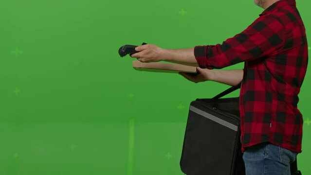 Caucasian male courier passing a box of pizza on green screen chroma key background. Man hold wireless bank payment POS terminal to process acquire credit card payment. Copy space mock up advertising