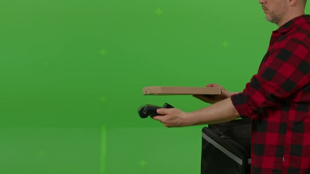 Caucasian male courier passing a box of pizza on green screen chroma key background. Man hold wireless bank payment POS terminal to process acquire credit card payment. Copy space mock up advertising