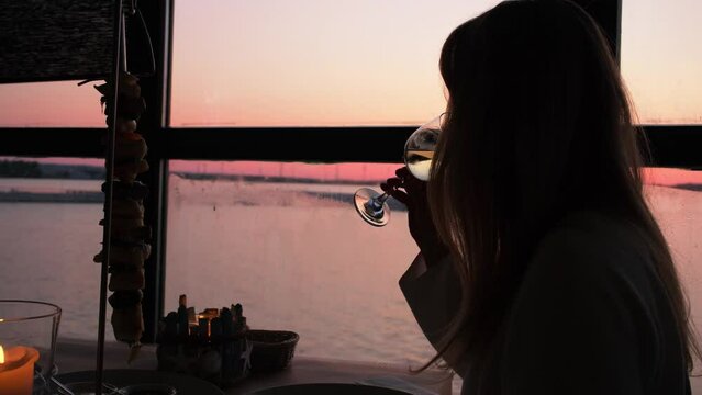 From behind the girl's shoulder, showcasing an exquisite table setting with wine glasses, flickering candles, and a hanging fish. The mesmerizing window view of the breathtaking sunset over the sea.