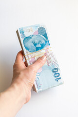 A woman's hand holds a stack of one hundred Turkish lira bills. A stack of one hundred Turkish lira banknotes tied with an elastic band in a hand on a light background.