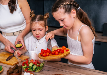 Obraz na płótnie Canvas A little girl is preparing a vegetarian salad and adding chopped tomatoes to a bowl together with her younger sister under the guidance of her mothe
