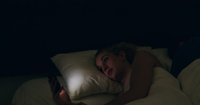 Phone, bed and night, woman online, insomnia and social media addiction with man sleeping and light in face. Cellphone, mobile app and couple in bedroom, girl reading post or meme on internet in dark
