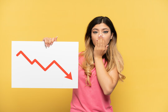 Young Russian girl isolated on yellow background holding a sign with a decreasing statistics arrow symbol with surprised expression