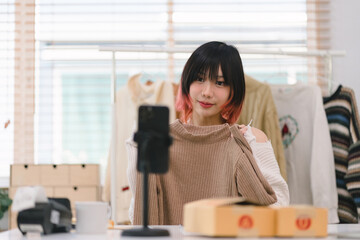 Young millennial Asian woman blogger showcasing clothes in front of a smartphone camera while recording a vlog video and live streaming from her shop.