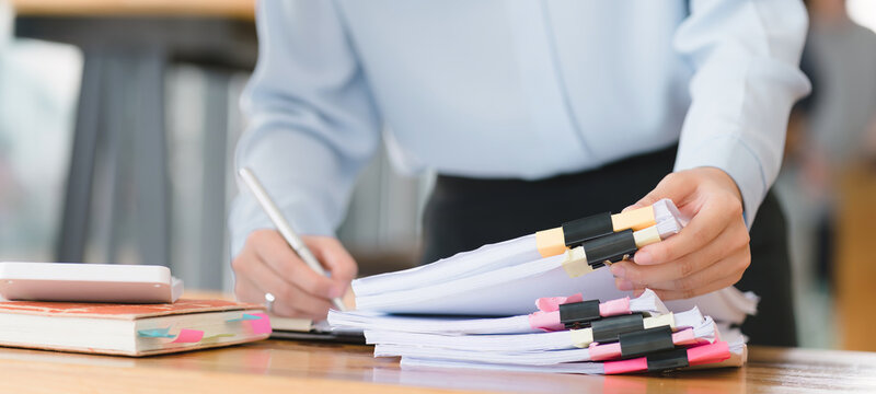 A businesswoman is sifting through stacks of paper files and folders that contain both incomplete and completed documents. Selective focus