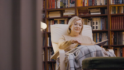 Relaxed senior woman covered in blanket reading a book at nursing home library