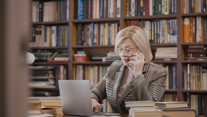 Busy woman in her 50s talking on the phone and working on her laptop in her home study