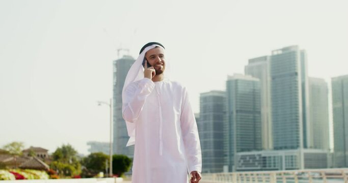 A young man in Arab national white clothes smiles, while talking on the phone walking along the embankment of a modern city on a sunny day