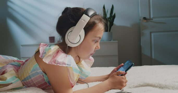 Cute Cheerful Schoolgirl in Headphones Uses a Smartphone While Lying on the Bed in the Bedroom in the Evening. Little Girl Watches Cartoons or Plays Games Using the Internet and Phone. 
