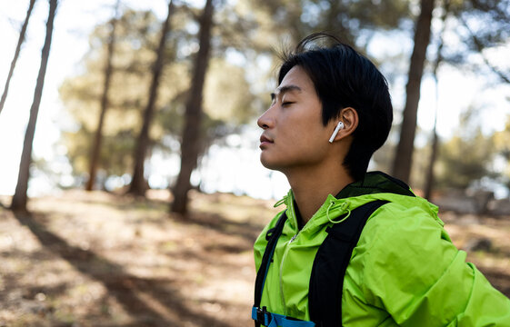 A young Asian man is hiking on top of a mountain. The young man enjoys the clean air of the countryside with his eyes closed. Concept of enjoying the environment, unwinding, relaxing outside the city.