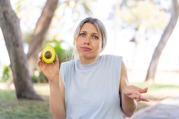 Young blonde woman holding an avocado at outdoors making doubts gesture while lifting the shoulders