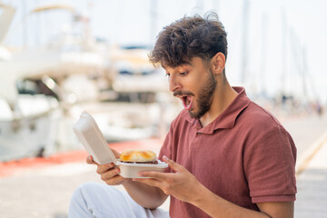 Young Arabian handsome man at outdoors holding a burger with surprised expression