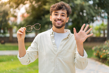 Young Arabian handsome man with glasses at outdoors saluting with hand with happy expression