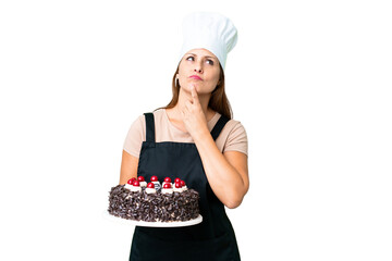 Middle age pastry chef woman holding a big cake over isolated background having doubts while looking up