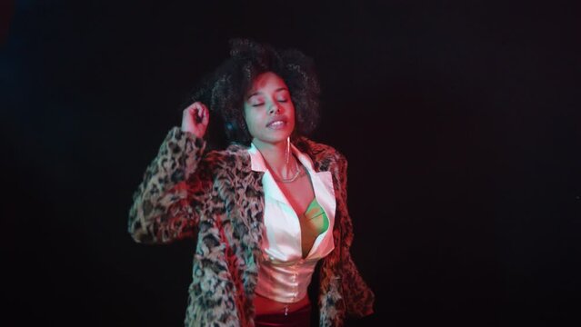 Young afro-haired and tattooed girl wearing a old-fashioned leopard fur coat, enthusiastically dancing on a black background while listening to music on headphones. (4/8)