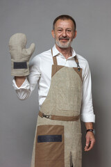 Mature man in a chef's apron waves, greets with an oven mitt and smiles.