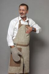A mature man in a chef's apron with an oven mitt stands and looks to the left.