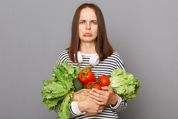 Green and organic for optimal health. Sad upset woman holding vegetables isolated over gray...