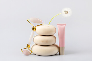 Facial anti-age massage for natural lifting and at home. Rose quartz facial roller and cosmetic bottle container on wooden stones with flower dandelion, gray background.