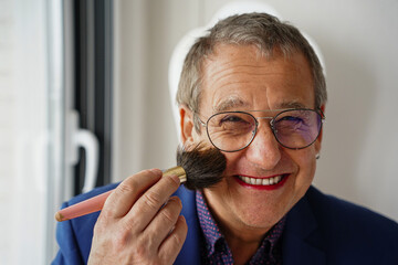 a portrait of an older man puts on makeup using the brush while looking at the camera. gay pride - obrazy, fototapety, plakaty