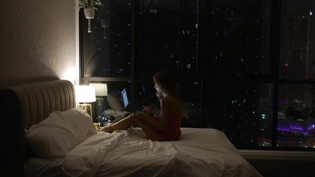 Young woman finish working on laptop and falling asleep in bedroom at night. Night city and skyscrapers in the background