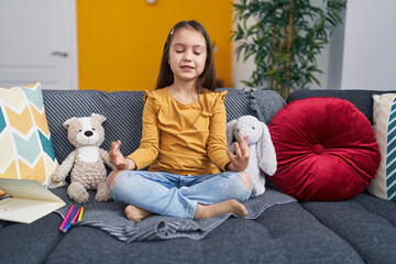 Adorable hispanic girl doing yoga exercise sitting on sofa at home