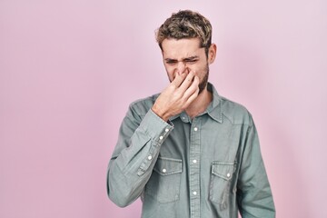 Hispanic man with beard standing over pink background smelling something stinky and disgusting, intolerable smell, holding breath with fingers on nose. bad smell