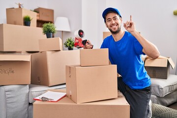 Hispanic man with beard working moving boxes surprised with an idea or question pointing finger with happy face, number one