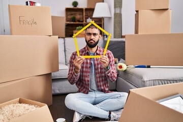 Middle east man with beard sitting on the floor at new home holding ruler skeptic and nervous,...