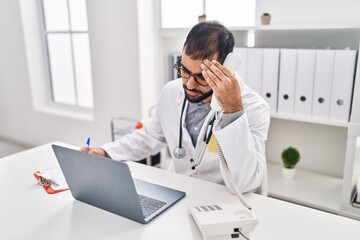 Young hispanic man doctor stressed writing medical report holding telephone at clinic