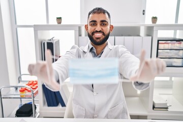 Young hispanic man doctor smiling confident holding medical mask at clinic