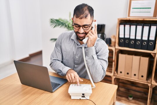 Young hispanic man business worker talking on telephone using laptop at office