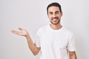 Handsome hispanic man standing over white background smiling cheerful presenting and pointing with...
